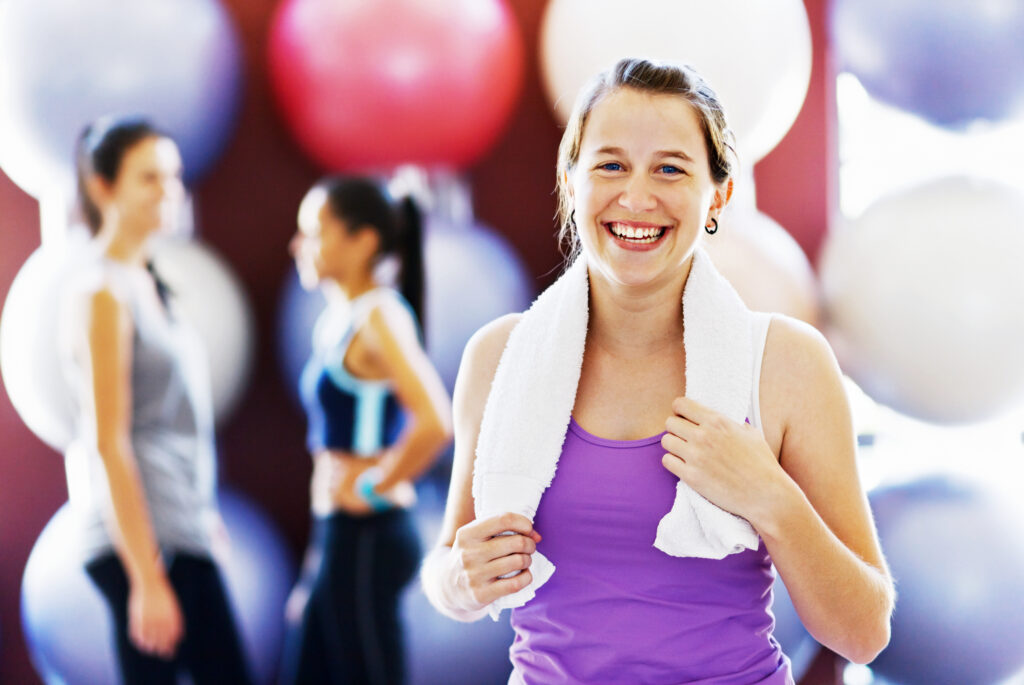 A pretty young woman smiles as she relaxes after an exercise session in the gym. More women stand in the background.